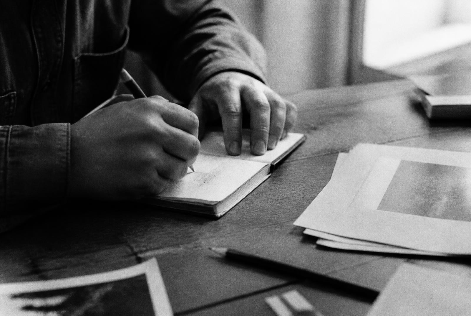 Hands writing in a notebook beside printed pages on a wooden table